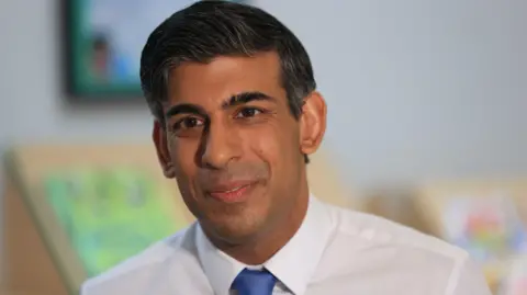 PA Media Rishi Sunak is smiling and wearing a white shirt and a blue tie