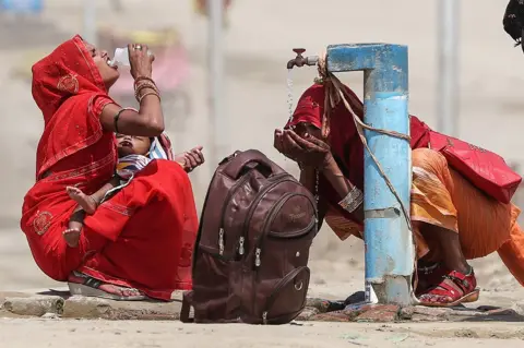 AFP Two women drink water and splash their face from a public tap on a street in Prayagraj, India, on 10 June. They both wear red, with one holding a baby and a backpack on the ground next to them.