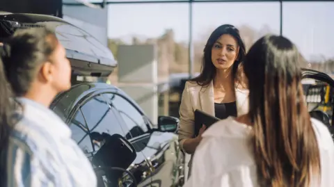 Getty Images A woman in a car dealership holding a tablet talks to two other people.