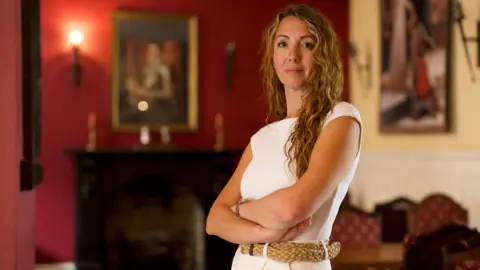 Samantha Harper, a woman with long curly blond hair, stands with her arms folded in the lounge of a historic pub with oil portraits hanging on the wall. 