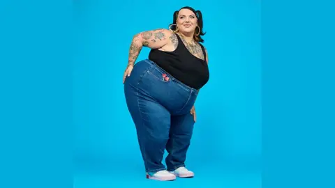 Snag Sophie who is a UK size 24 to 26 model on a shoot wearing blue jeans and a black vest top and glamorous make up, posing with her hand on one hip against a bright blue background. she has tattoos on her arm and chest.