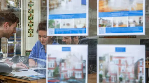 Getty Images Couple seen looking at paperwork through the window of an estate agent with adverts seen on the window.