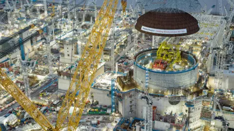 EDF An evening aerial view - a crane lowers the dome of a nuclear reactor on to a building. The site is surrounded by cranes, scaffolding and construction. This is Hinkley Point C, the nuclear power station being built in Somerset. 