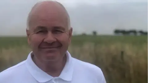 A head-and-shoulders shot of Neil Lockhart in a buttoned-up white polo shirt, stood in front of grassland and a field with trees silhouetted on the horizon under a grey sky.