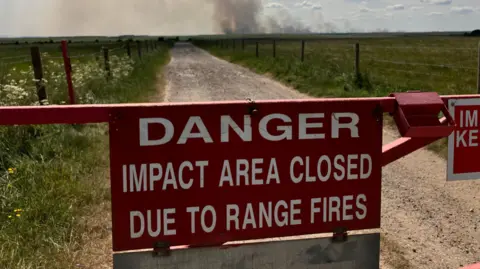 BBC A large red warning sign sits on a red barrier across a fenced dirt track that runs through a countryside plain. Large white writing warns "Danger. Impact area closed due to range fires". In the distance a cloud of smoke rises into the sky.