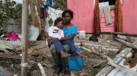 Reuters A mother and daughter sit outside their home with fallen tree branches and other debris surrounding them. The mother is wearing a blue top and the daughter a white Nike jumper. 