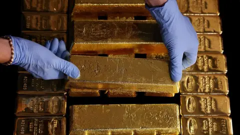 Bloomberg via Getty Images A pair of light blue latex-gloved hands place a gold bar on top of pile of other gold bars. The person has a gold bracelet on their right wrist.