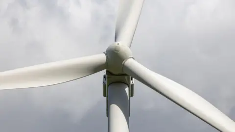 Getty Images Close-up of a wind turbine’s rotor and three blades against a cloudy sky, viewed from below.