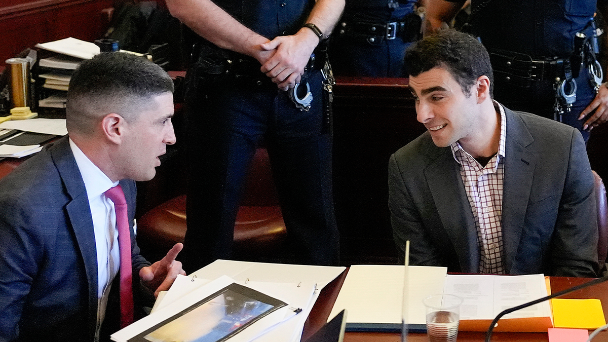 Luigi Mangione in court wearing a checkered shirt and sportsjacket while leaning over to speak with a member of his legal team
